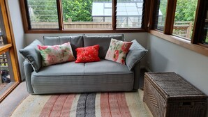 A grey sofa with floral cushions, a patterned rug, and a wicker basket in a room with large windows. - Lark Cottage - relaxing retreat (Burrawang)