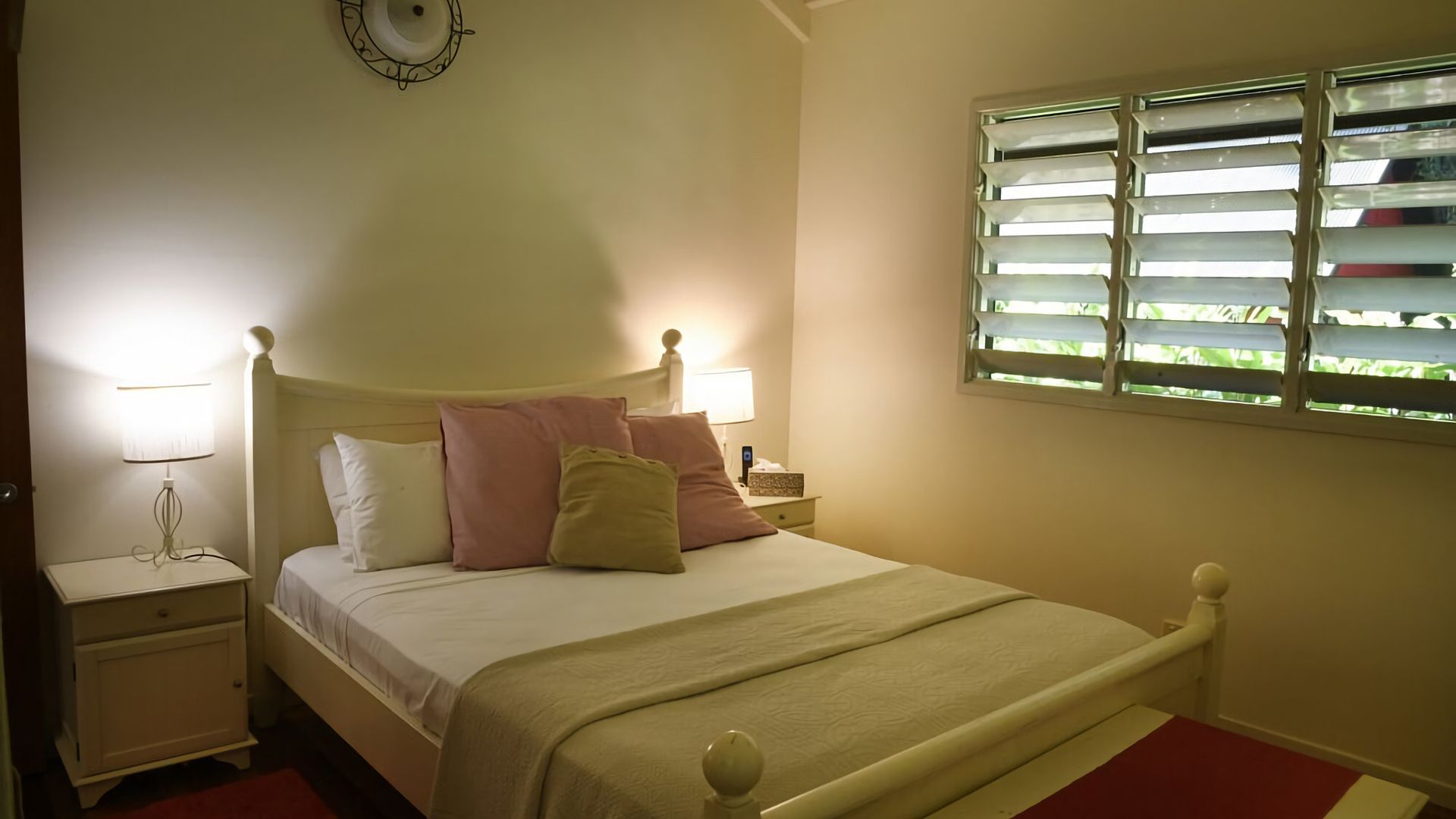 A bedroom with a bed, bedside lamps, a nightstand, a window with blinds, and a wall clock.