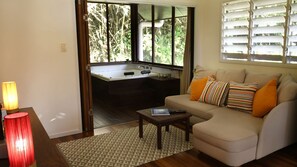 Un salon comprenant un canapé, une table basse, une lampe et donnant sur une cuisine grâce à une porte vitrée.