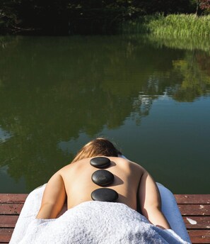 Une personne reçoit un massage du dos avec trois pierres posées sur son dos, assise sur une jetée en bois au bord d’une étendue d’eau calme.