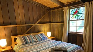 A wooden-paneled room with a bed, two bedside lamps, and a window with a stained glass bird design. - Willow Cottage @ Hidden Valley Retreat Cottages (Grose Vale)