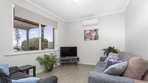 Uma sala de estar moderna com uma janela grande, uma televisão de tela plana, um sofá cinza, uma mesa pequena e um vaso com planta.