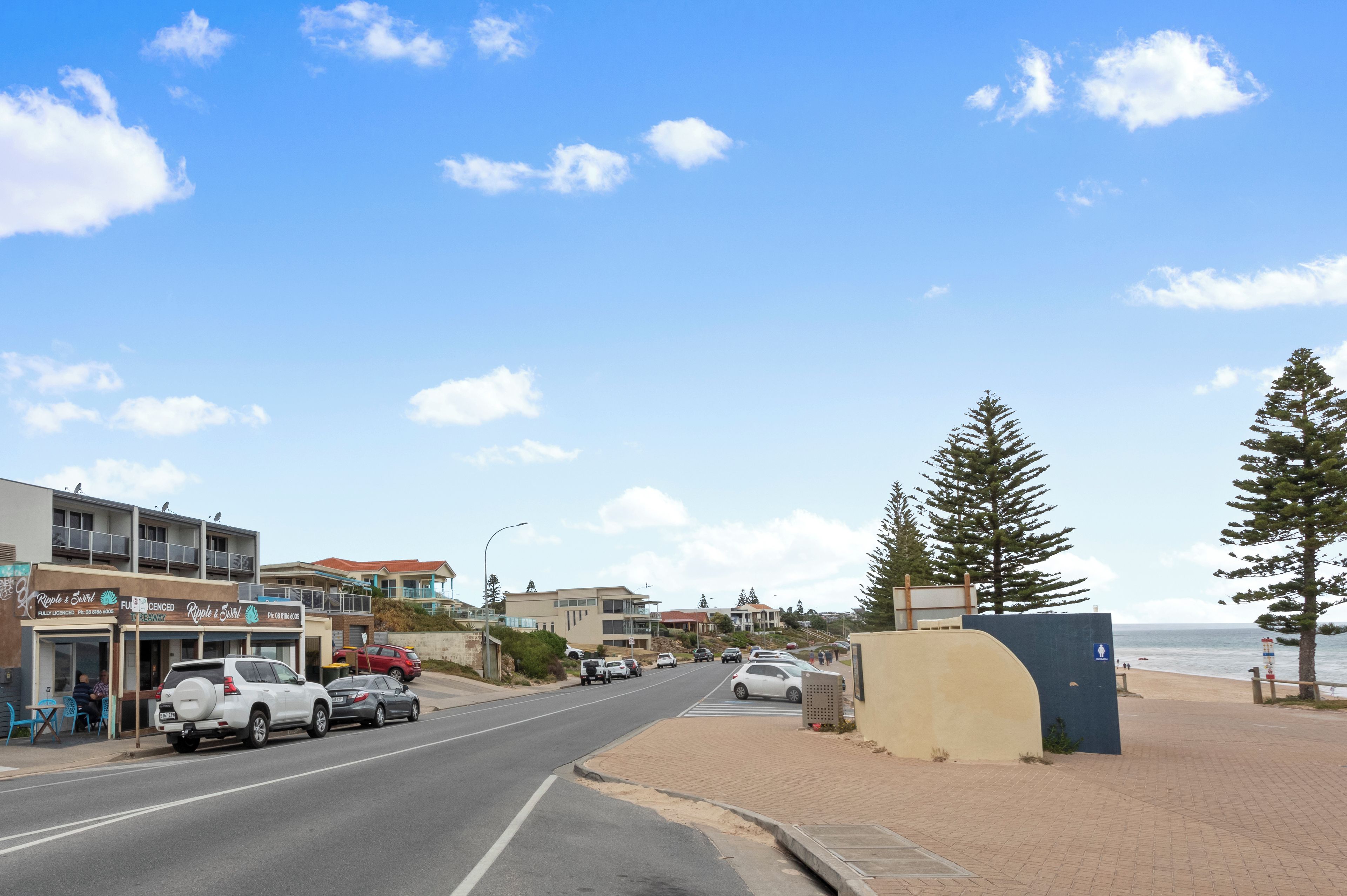A coastal street with parked cars, a beach, and buildings with signage.
