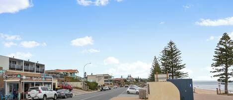 A coastal street with parked cars, a beach, and buildings with signage.