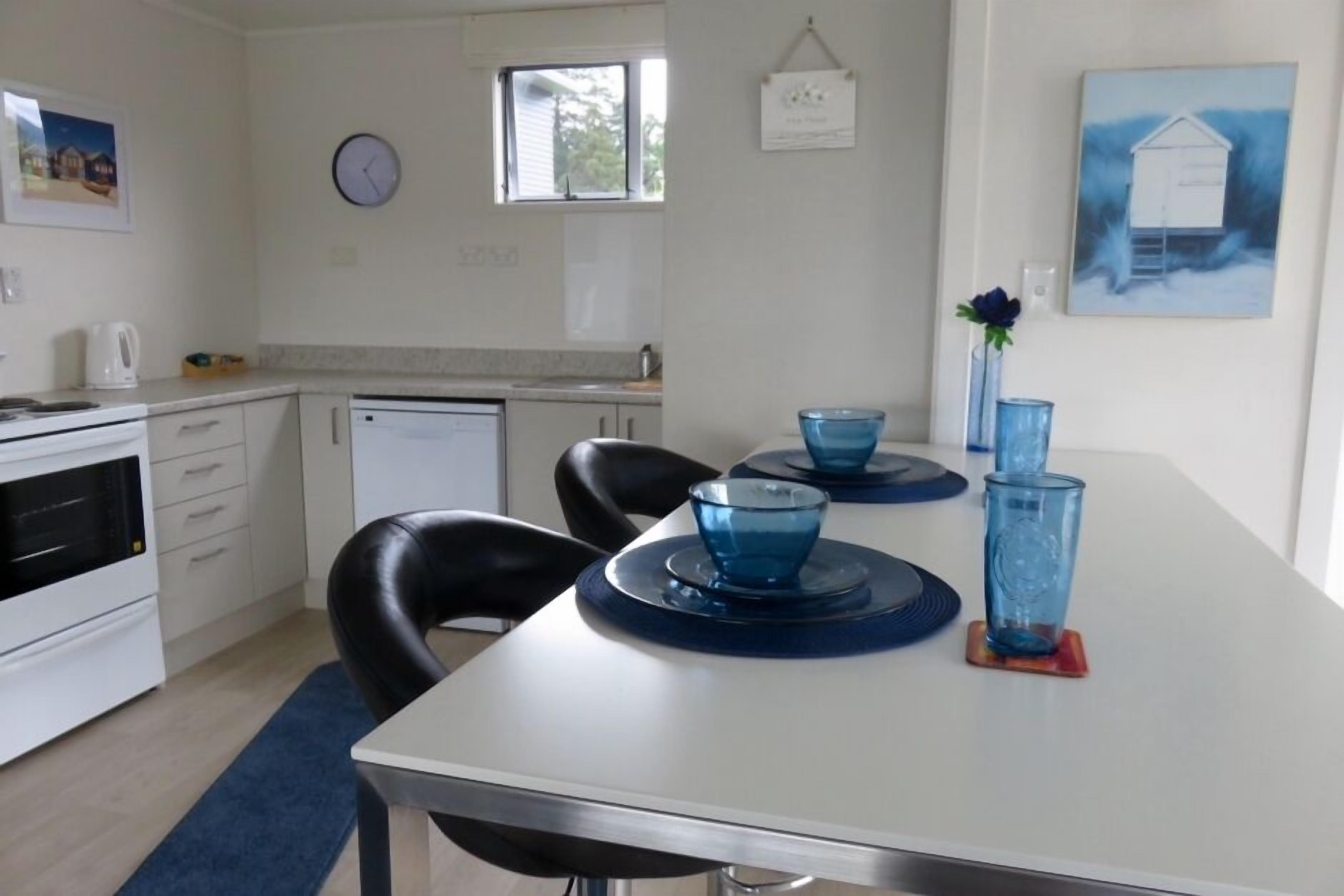 Una cocina moderna con armarios blancos, una estufa blanca y una silla negra. Hay una mesa de comedor con cristalería azul y un florero con una flor.