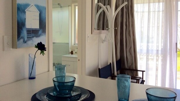 A dining area with a white table, blue glassware, and a painting of a house. - Garden Studio ~ Coromandel Apartments (Coromandel Township)