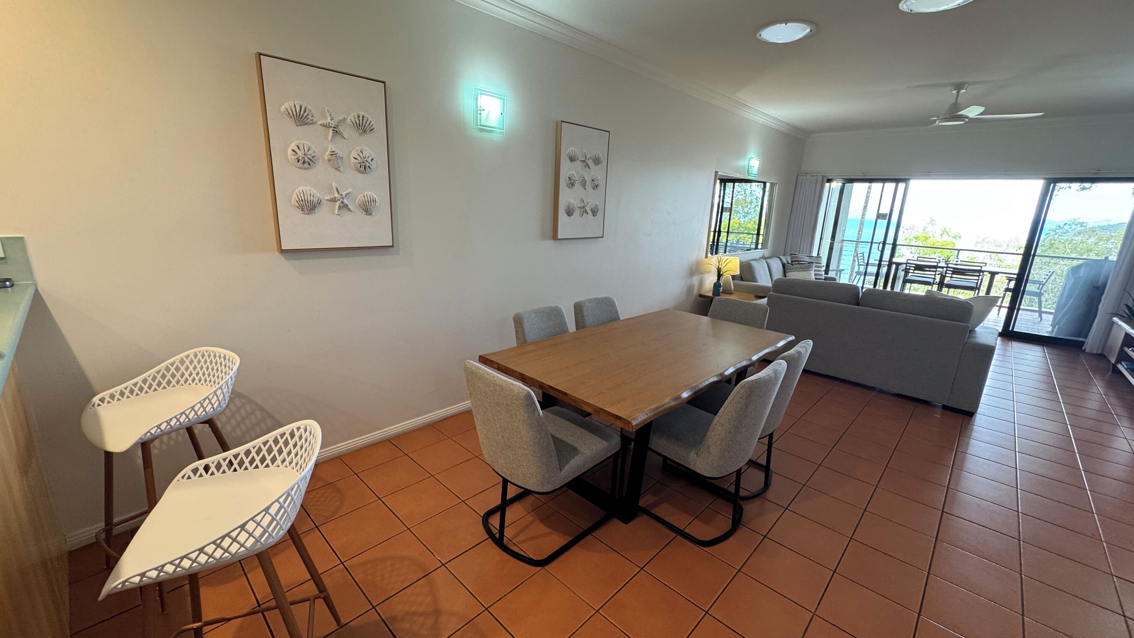 A spacious living area with a wooden dining table, grey upholstered chairs, a grey sofa, and a balcony with outdoor furniture.