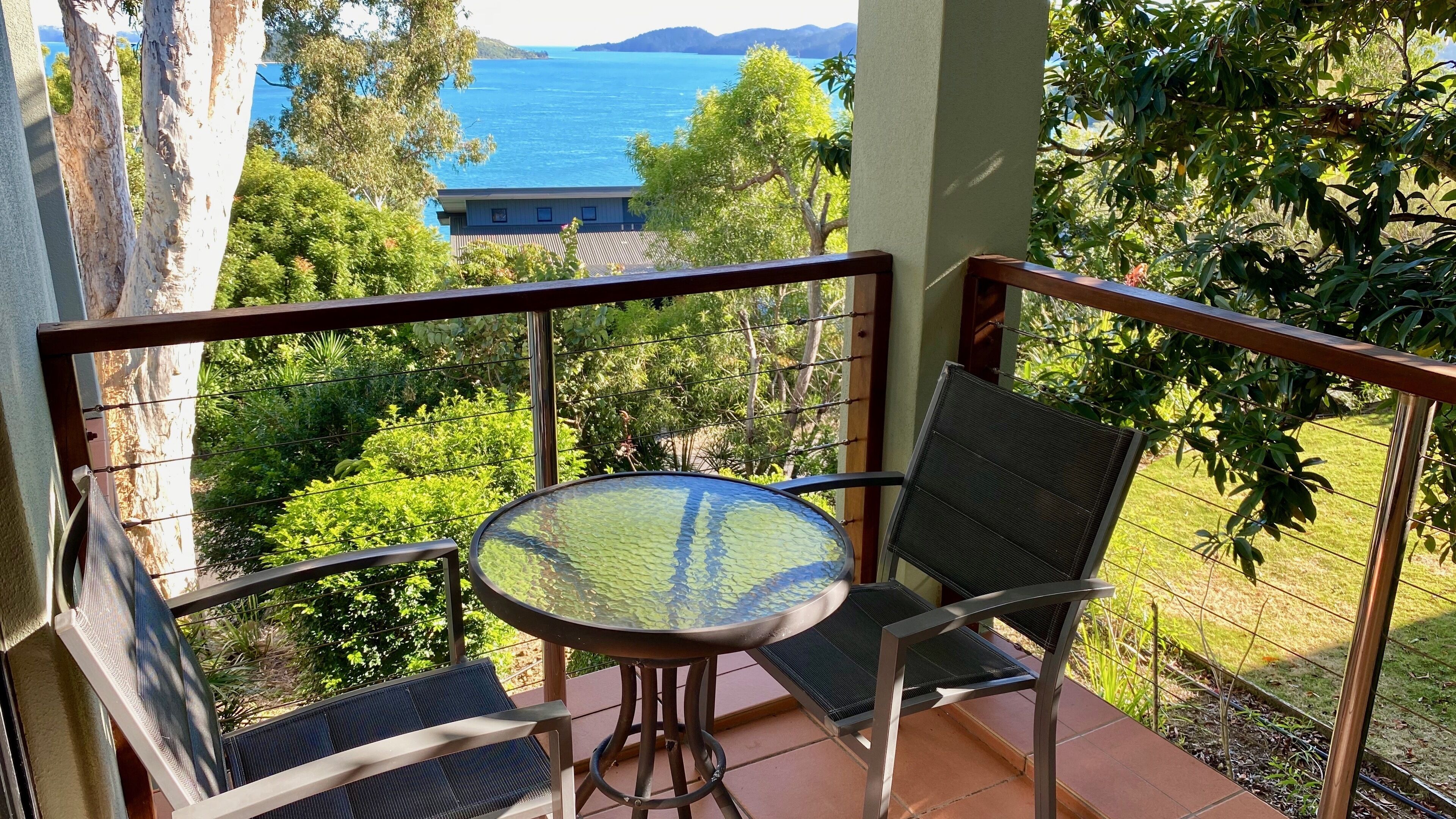 Un balcon avec une table et des chaises, offrant une vue panoramique sur la mer et la verdure.