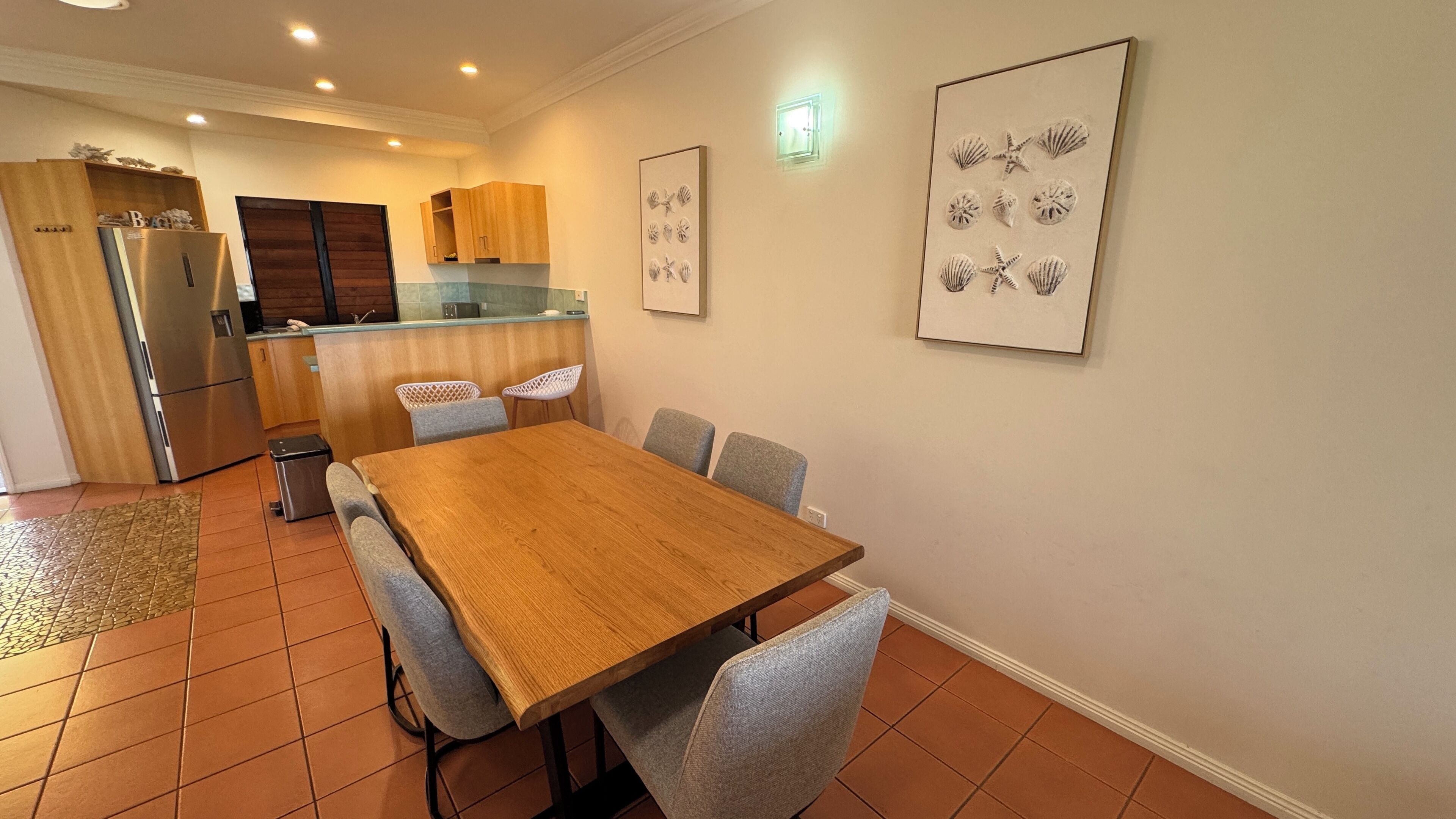 A dining area with a wooden table and grey chairs, a kitchen with wooden cabinets and a stainless steel refrigerator, and framed artwork on the walls.