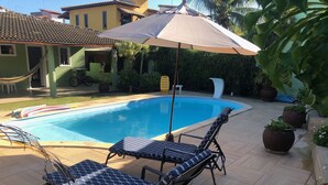 Una piscina con sillas de descanso y una sombrilla, rodeada de plantas en macetas y un patio con baldosas.