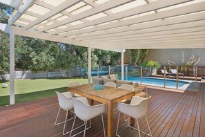 Un patio exterior con mesa y sillas de madera, una piscina y una zona cubierta con vista al jardín trasero.