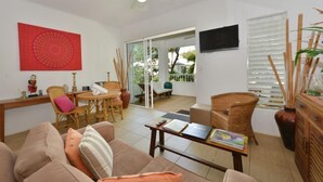 Ein Wohnzimmer mit Sofa, Sesseln, Tisch und Fernseher. Eine Glastür führt in einen Außenbereich.