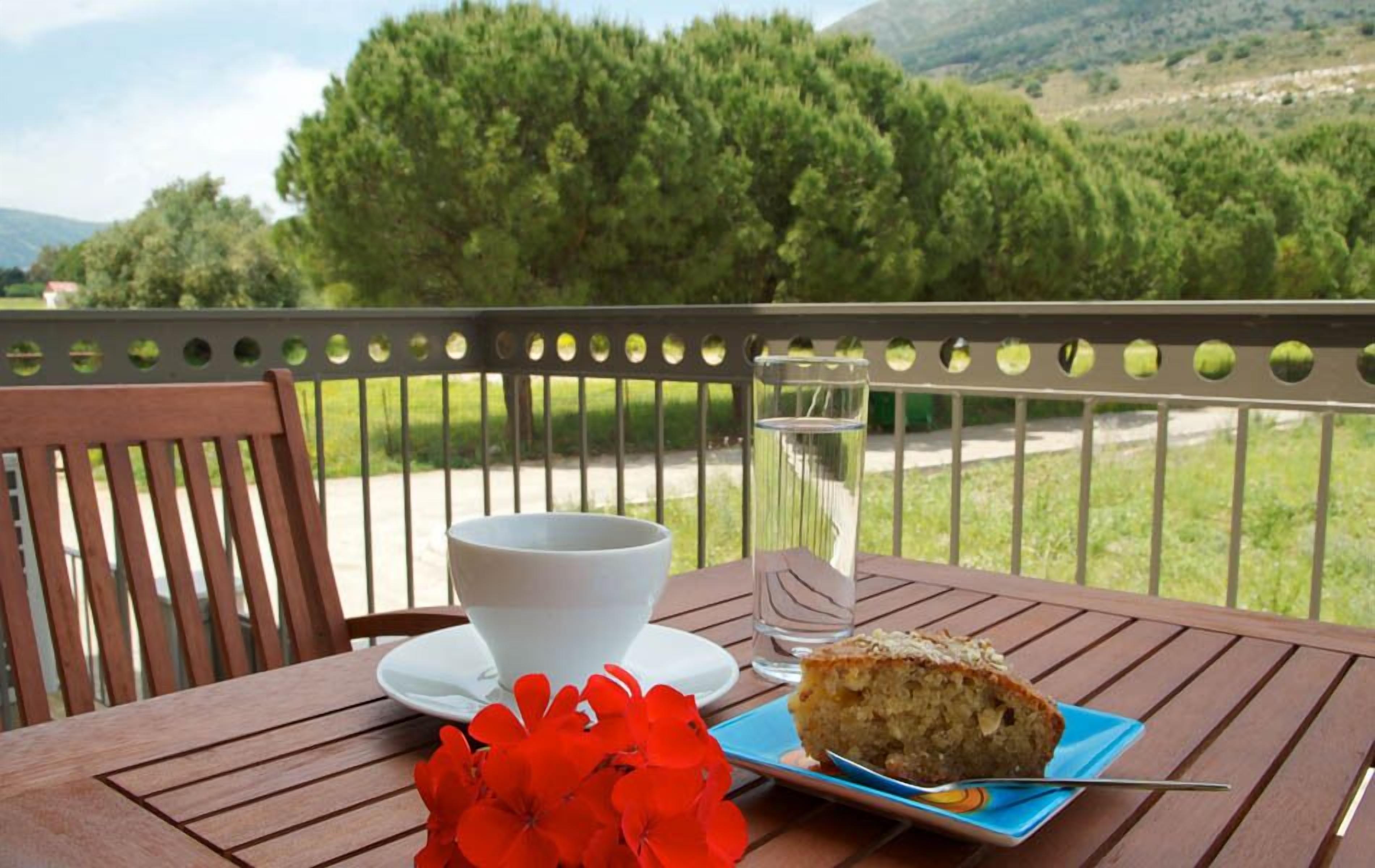 Una mesa con una taza blanca, un vaso de agua y una porción de pastel sobre un plato azul, ubicada en una terraza de madera con vista a árboles y colinas verdes.
