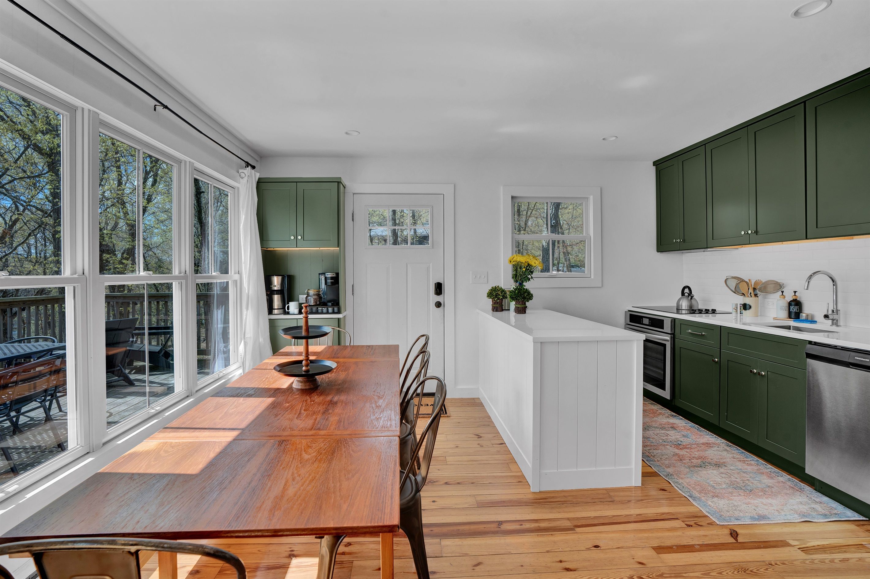 A modern kitchen with a wooden dining table, green cabinets, and large windows.