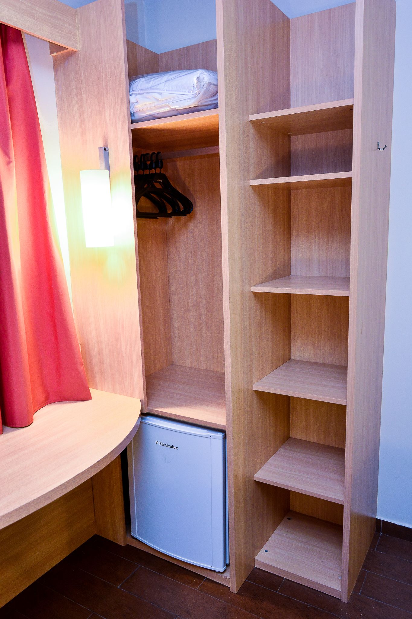 A compact room with a wooden wardrobe, a small refrigerator, and a red curtain.
