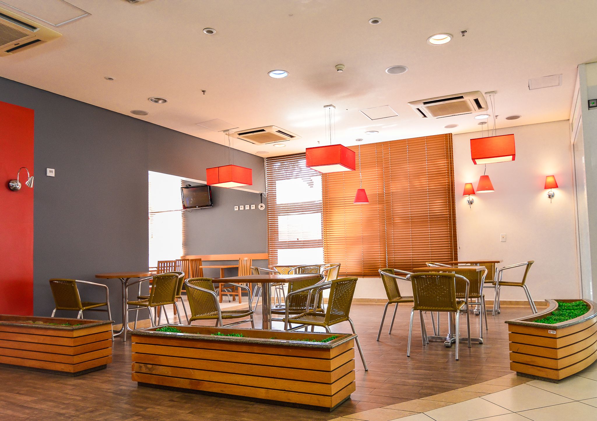 A modern indoor space with wooden flooring, metal chairs, and tables. There are built-in planters with green plants and recessed ceiling lighting.