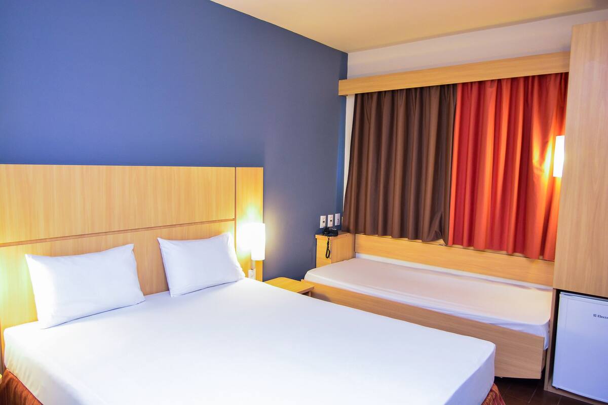 A hotel room with a large bed, bedside tables, a nightstand, a lamp, a window with curtains, and a television.