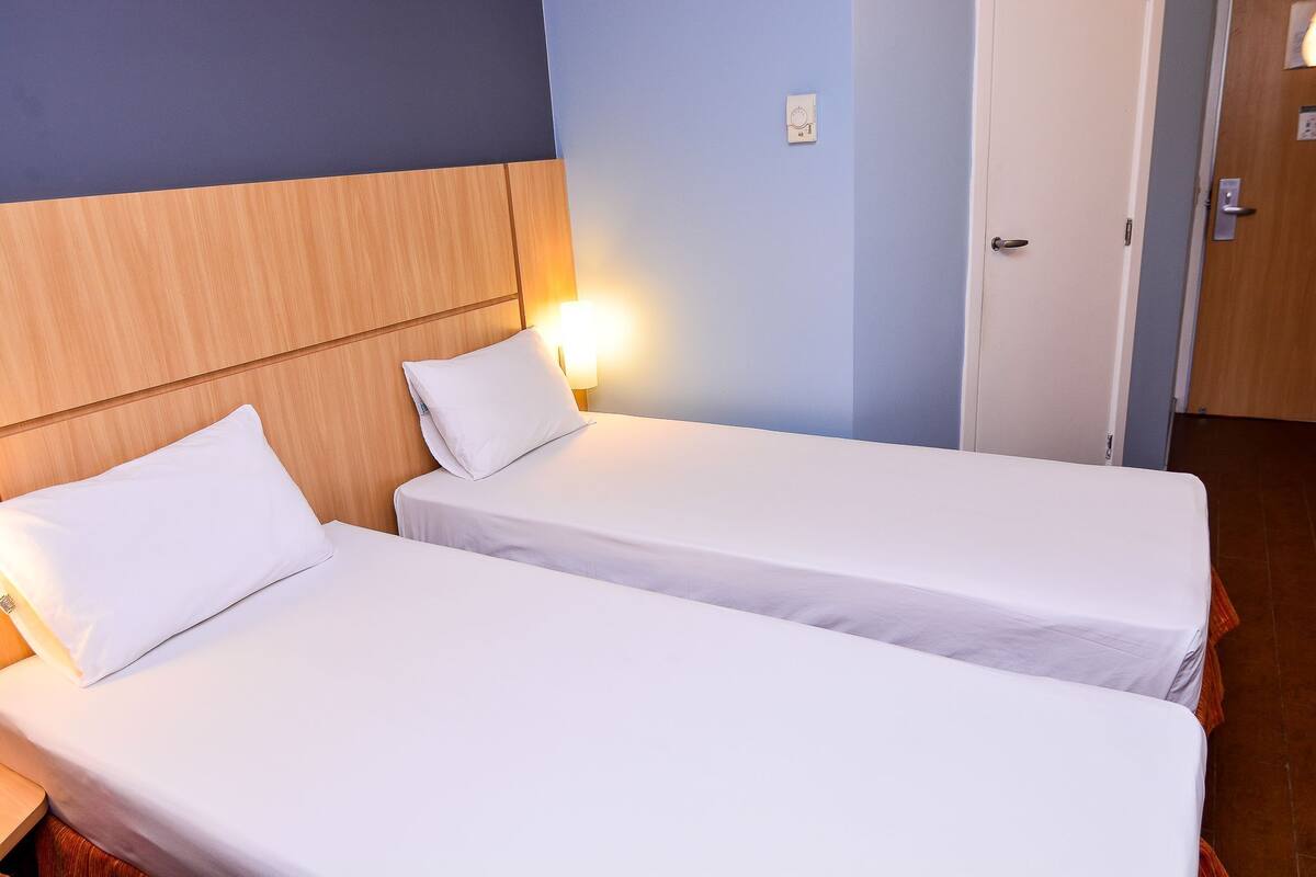 Two single beds with white linens, a wooden headboard, a wall-mounted light, and a door.