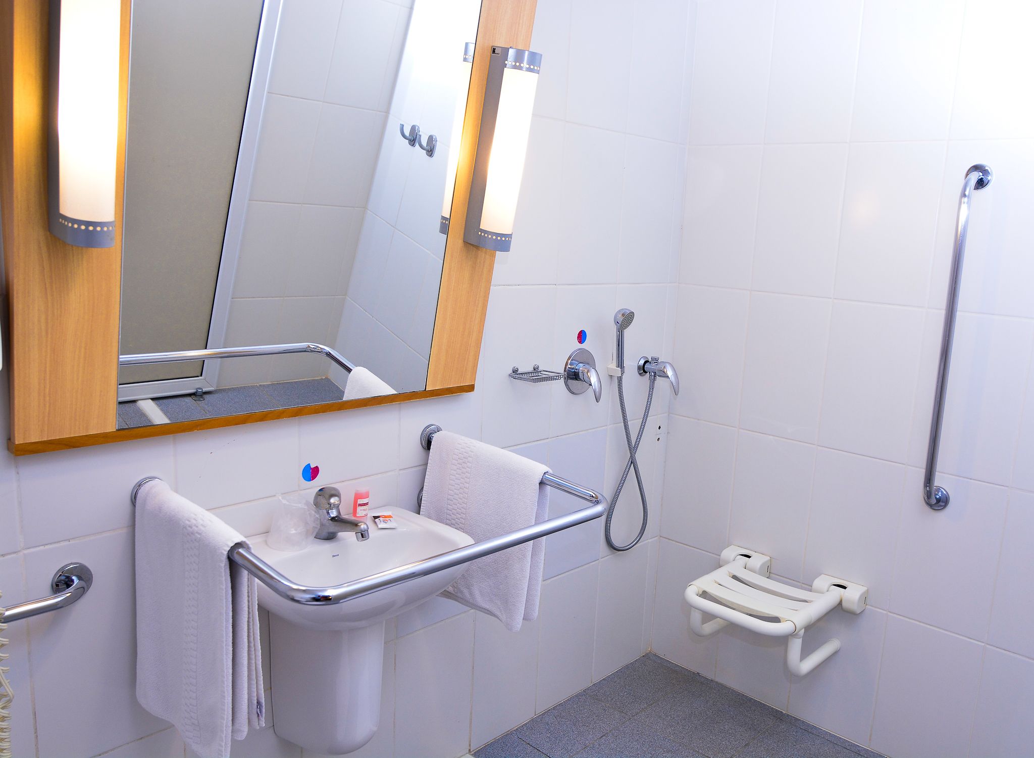 Un baño con lavabo, espejo, toallero, barra de apoyo y mandos de ducha.