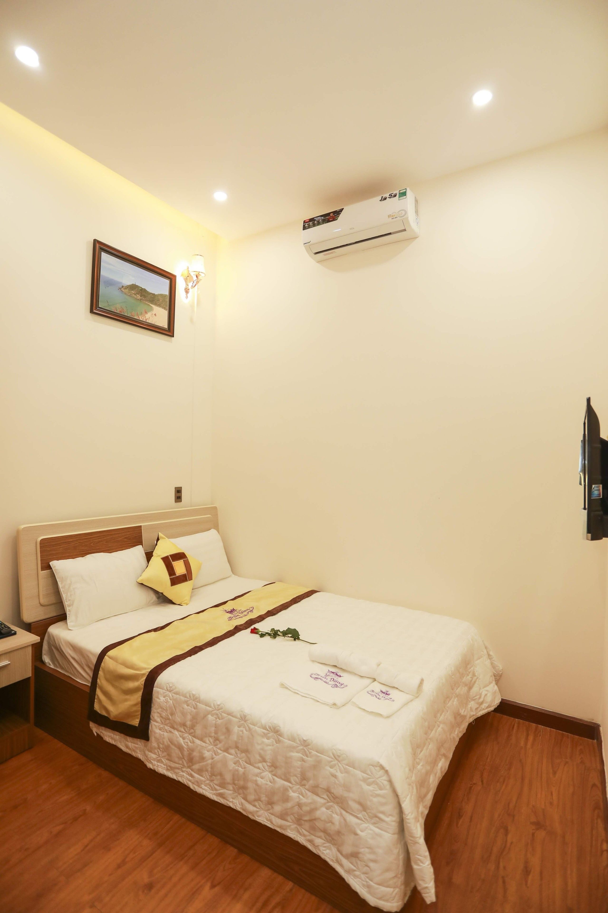 A neatly made bed with a white comforter and a brown headboard, a bedside table with a lamp, and an air conditioning unit on the wall.