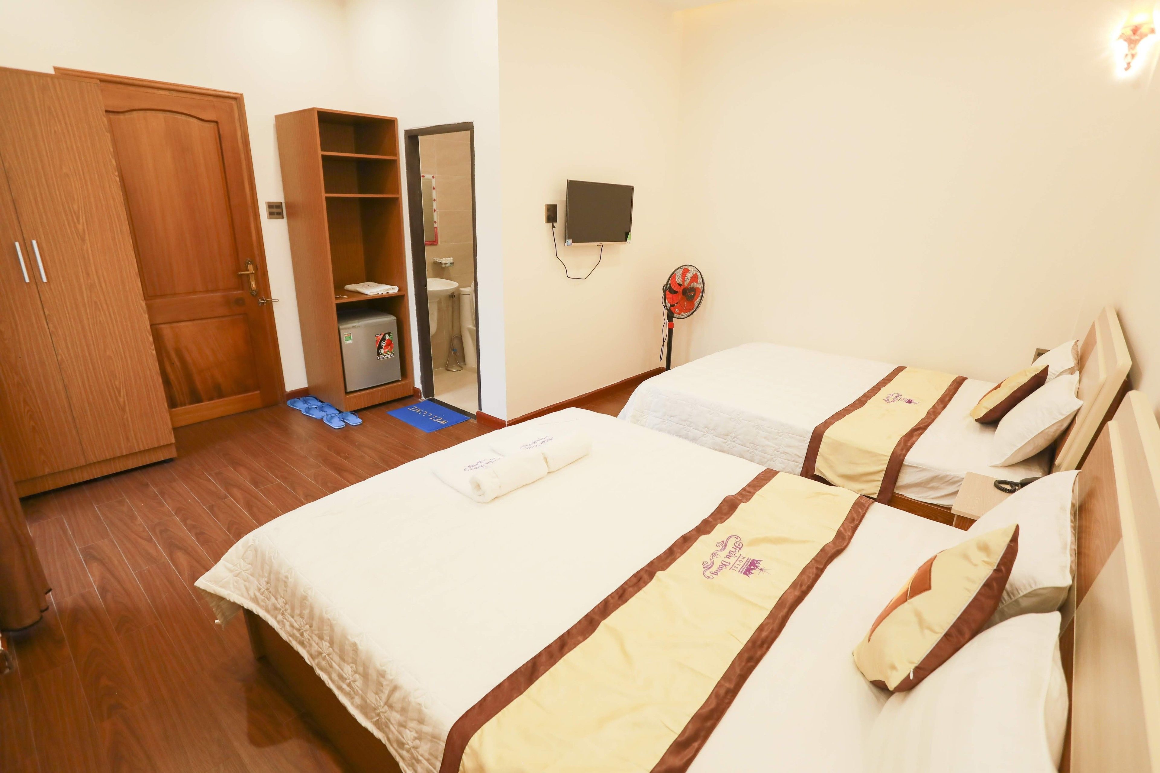 A hotel room with two beds, a wardrobe, a mirror, a small shelf, a wall-mounted TV, and a fan.
