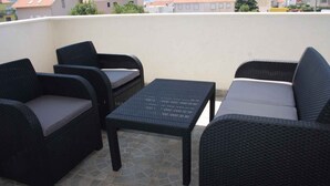 A terrace with wicker furniture, including two chairs and a table, set against a backdrop of buildings and a mountain.