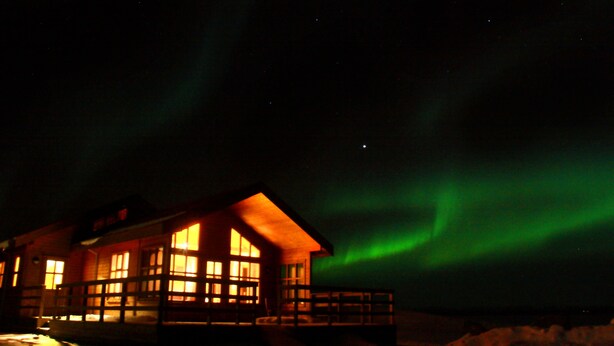A wooden cabin with multiple windows, surrounded by snow, under a night sky with green aurora. - Vorsabær 2 Holiday Home (Selfoss)