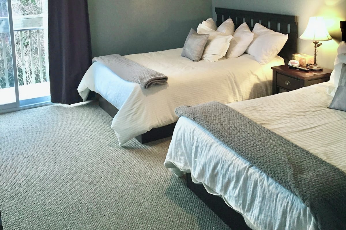 Habitación de hotel con dos camas, un ventilador de techo, una mesita de noche con lámpara y vistas al exterior a través de una ventana con cortinas.