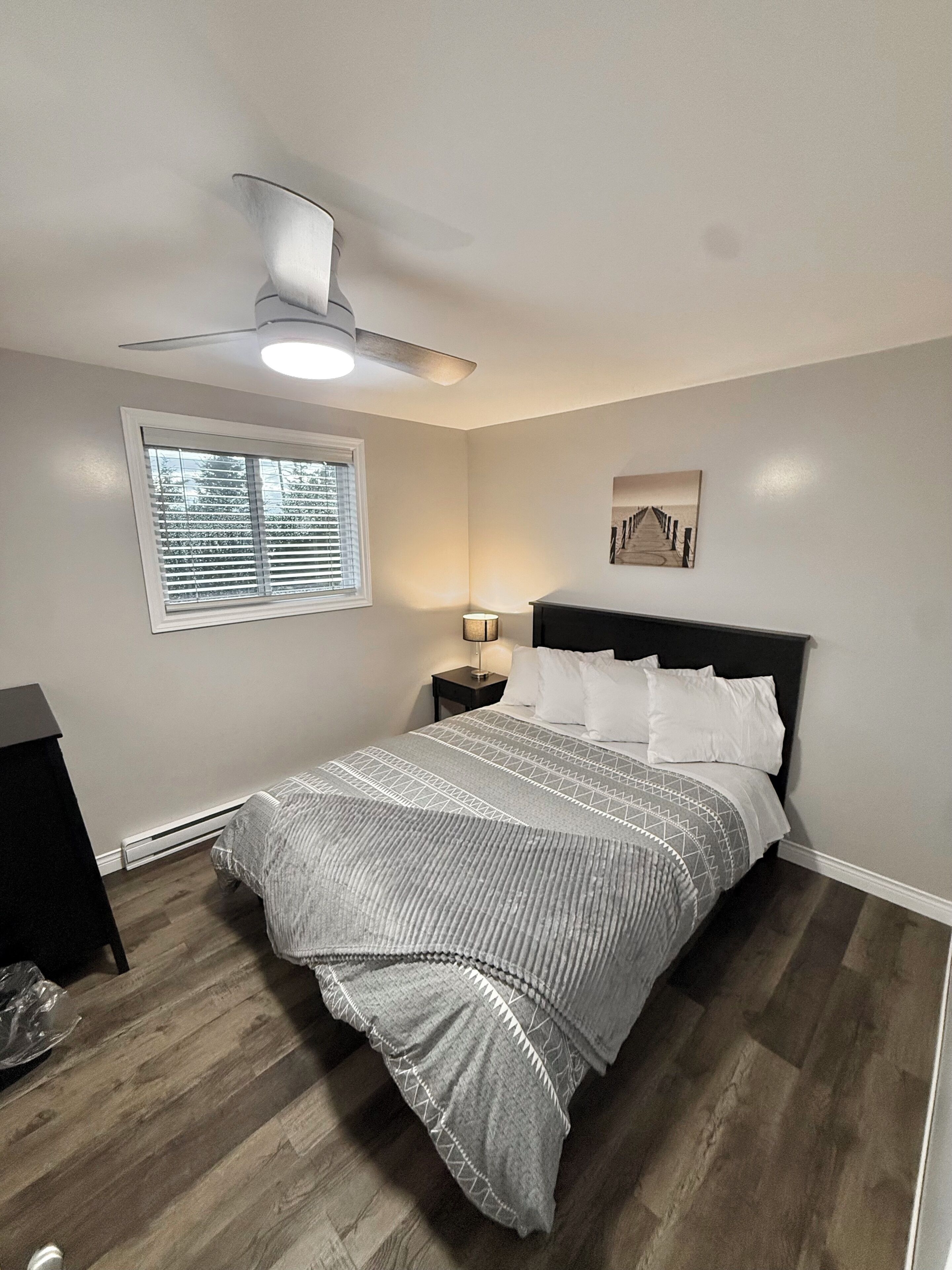 A bedroom with a bed, a nightstand, a ceiling fan, a window with blinds, and a wall-mounted picture.