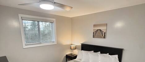 A bedroom with a bed, a nightstand, a ceiling fan, a window with blinds, and a wall-mounted picture.