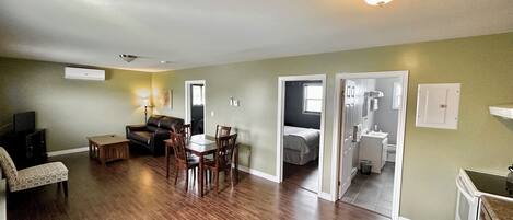 A spacious living room with hardwood floors, a dining area with a table and chairs, a sofa, and a TV. There is a door leading to a bedroom and a bathroom.