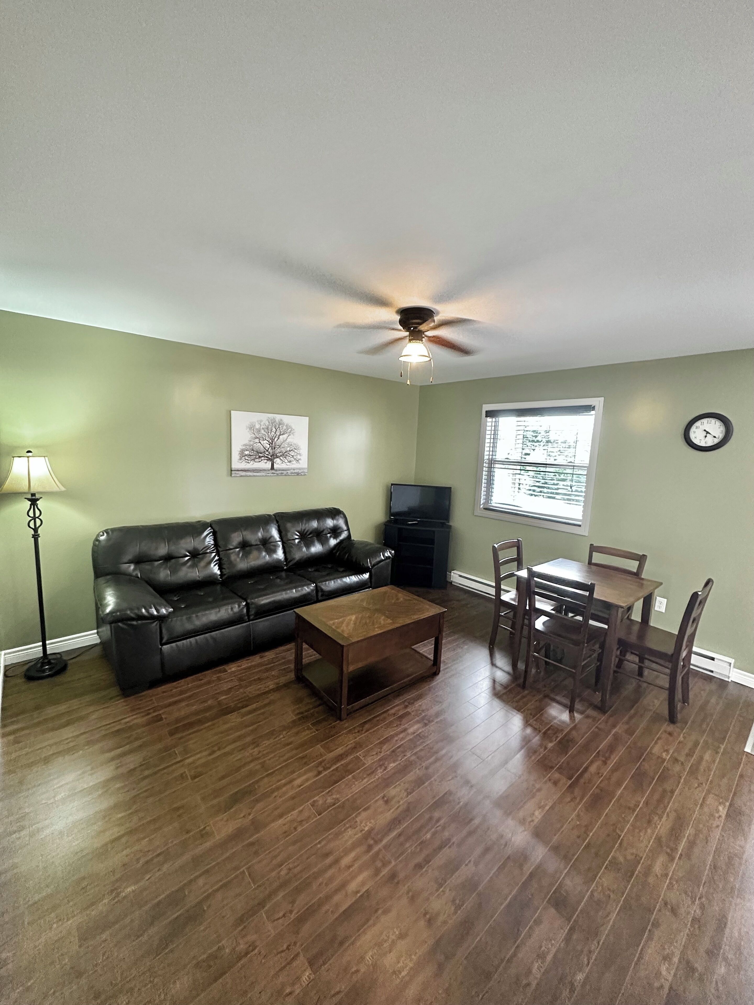 A living room with a black leather sofa, a wooden coffee table, a dining table with chairs, a ceiling fan, a TV, and a clock on the wall.