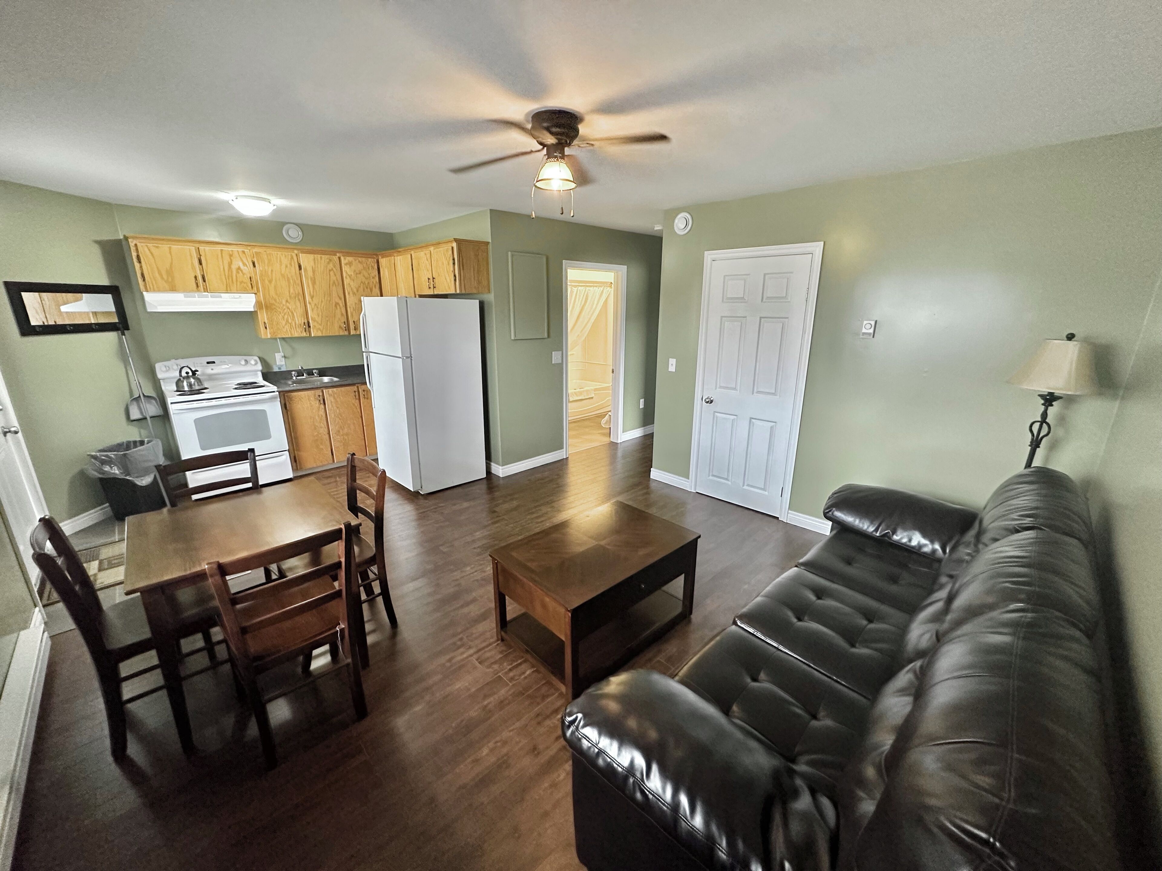 A spacious living area with a leather sofa, wooden dining table, and chairs. A kitchen with wooden cabinets and a refrigerator is visible in the background.
