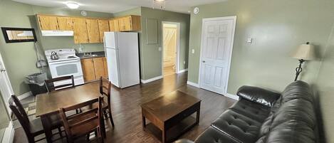 A spacious living area with a leather sofa, wooden dining table, and chairs. A kitchen with wooden cabinets and a refrigerator is visible in the background.