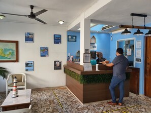 Un espace d’accueil d’hôtel avec un comptoir de réception en bois, un ventilateur de plafond et divers affiches d’information accrochées aux murs.