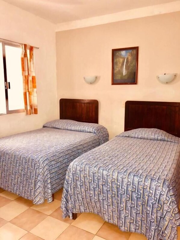 A room with two single beds, a window with curtains, a painting on the wall, and a wall-mounted light fixture.
