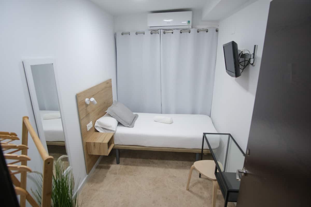 A small, modern bedroom with a single bed, a wooden headboard, a bedside table with a lamp, a wall-mounted TV, and a large window with curtains.