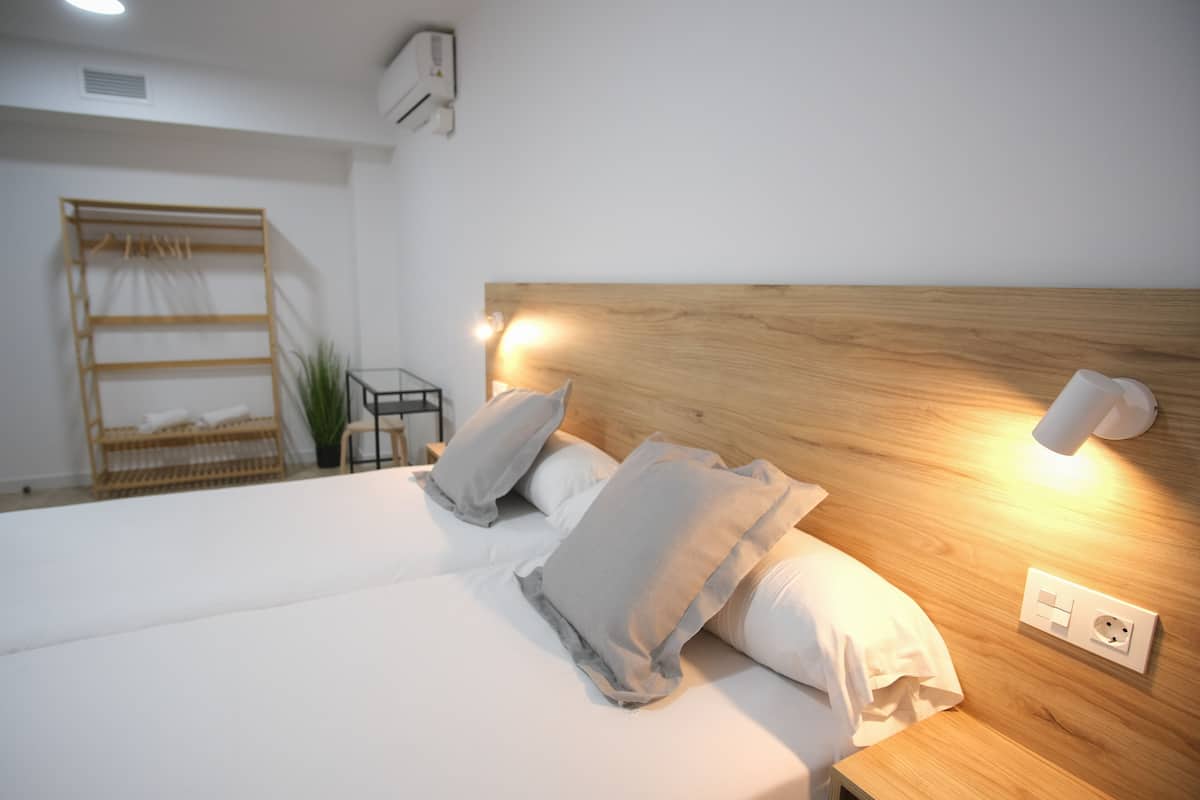 A neatly made bed with pillows, a bedside table, a wall-mounted light, and a power outlet.