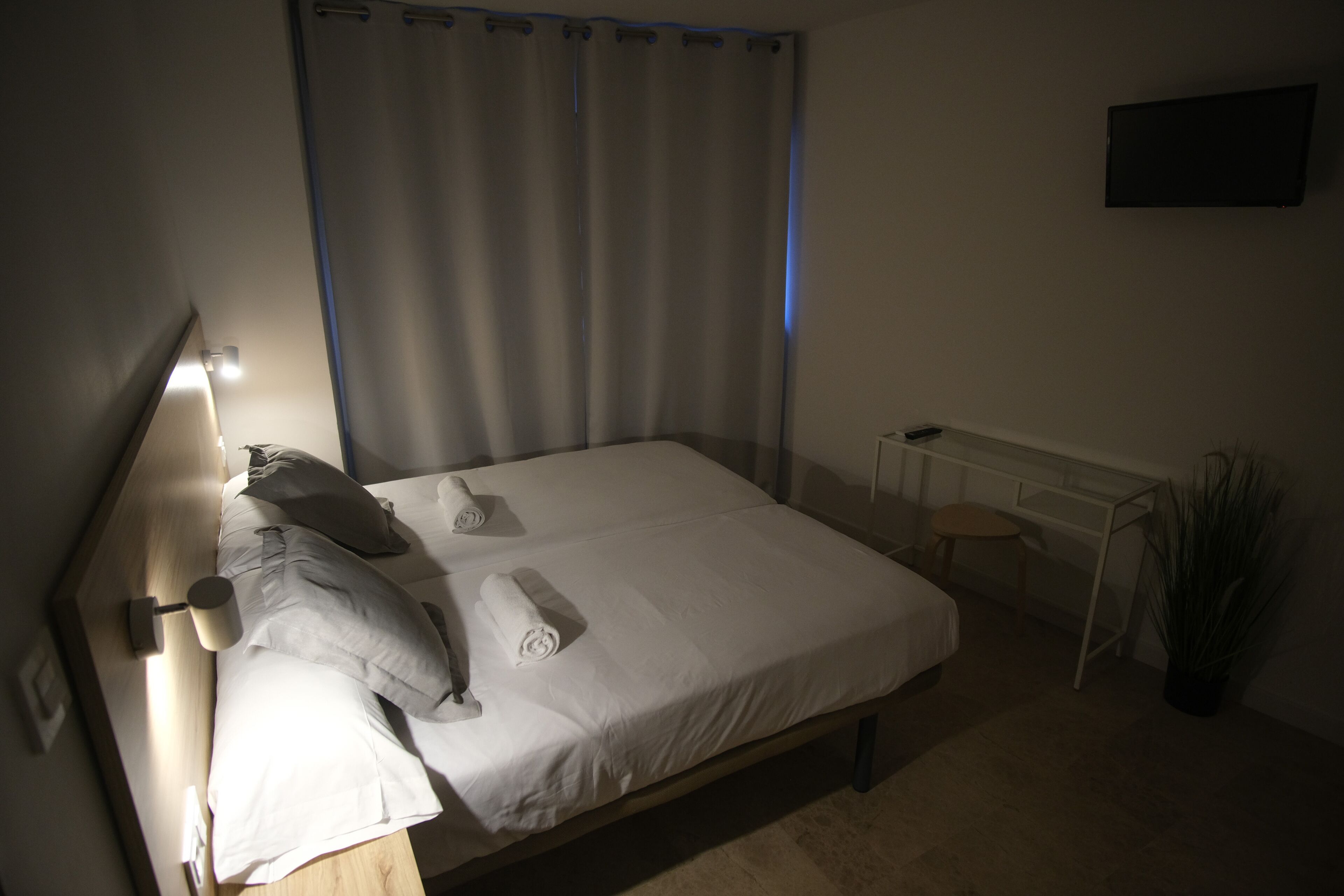 En pænt redt seng med hvide sengetøj og puder, et natbord med lampe, en lille skammel og en potteplante.