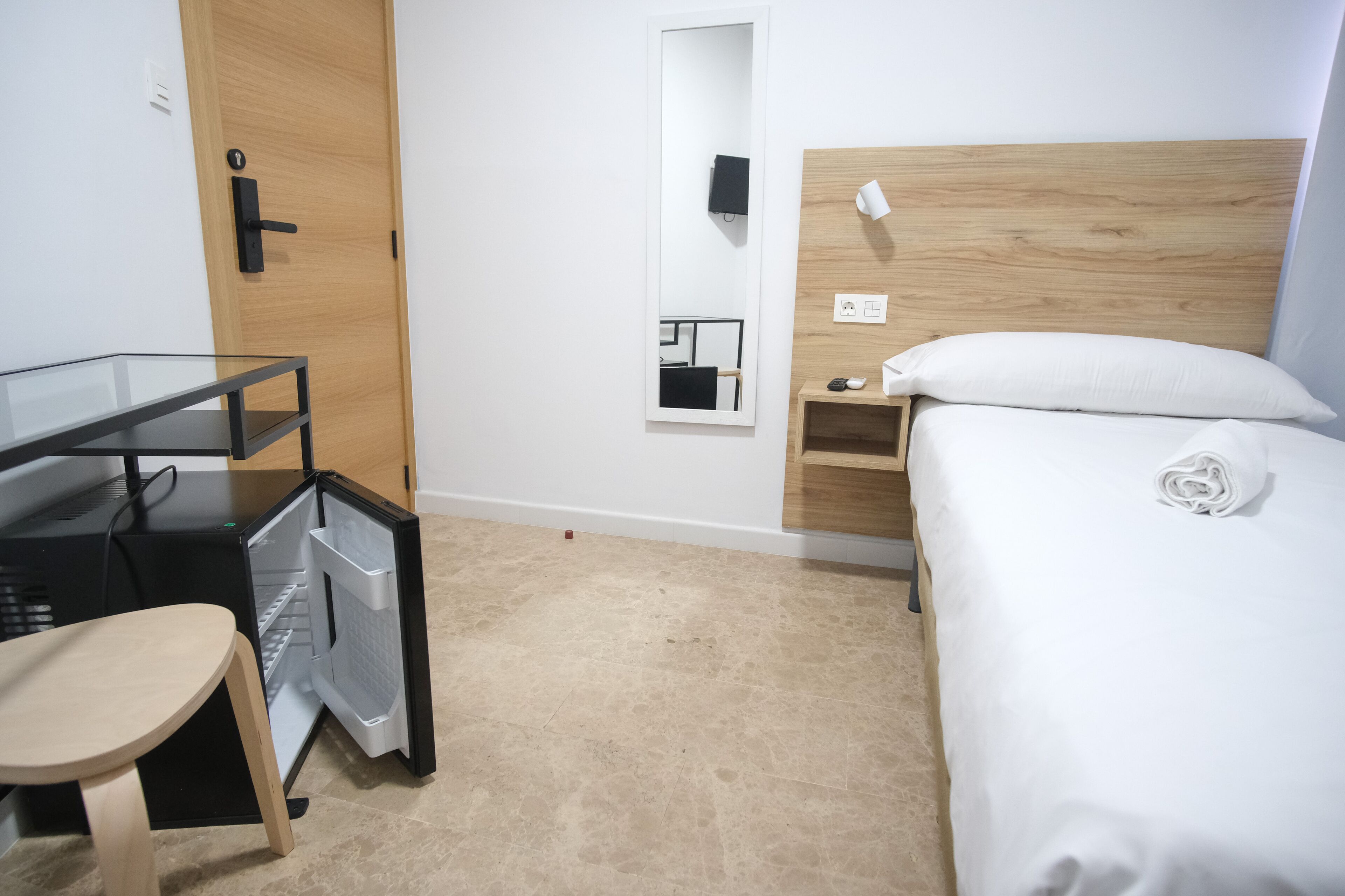 Hotellihuone, jossa on sänky, työpöytä tietokoneella, tuoli, peili ja yöpöytä lampun kanssa.