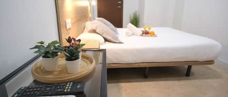 A hotel room with a bed, a bedside table with a remote control, a tray with plants, and a wall-mounted lamp.