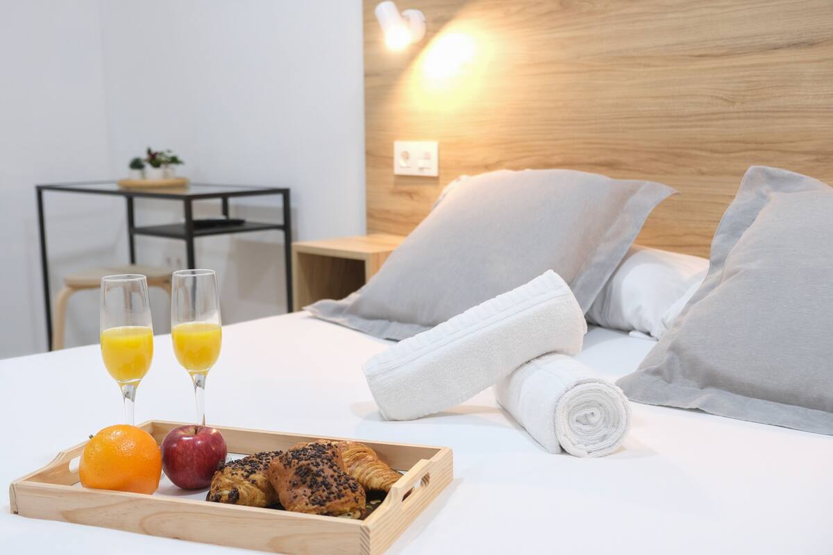 A neatly made bed with pillows, a rolled towel, and a tray with food and drinks.
