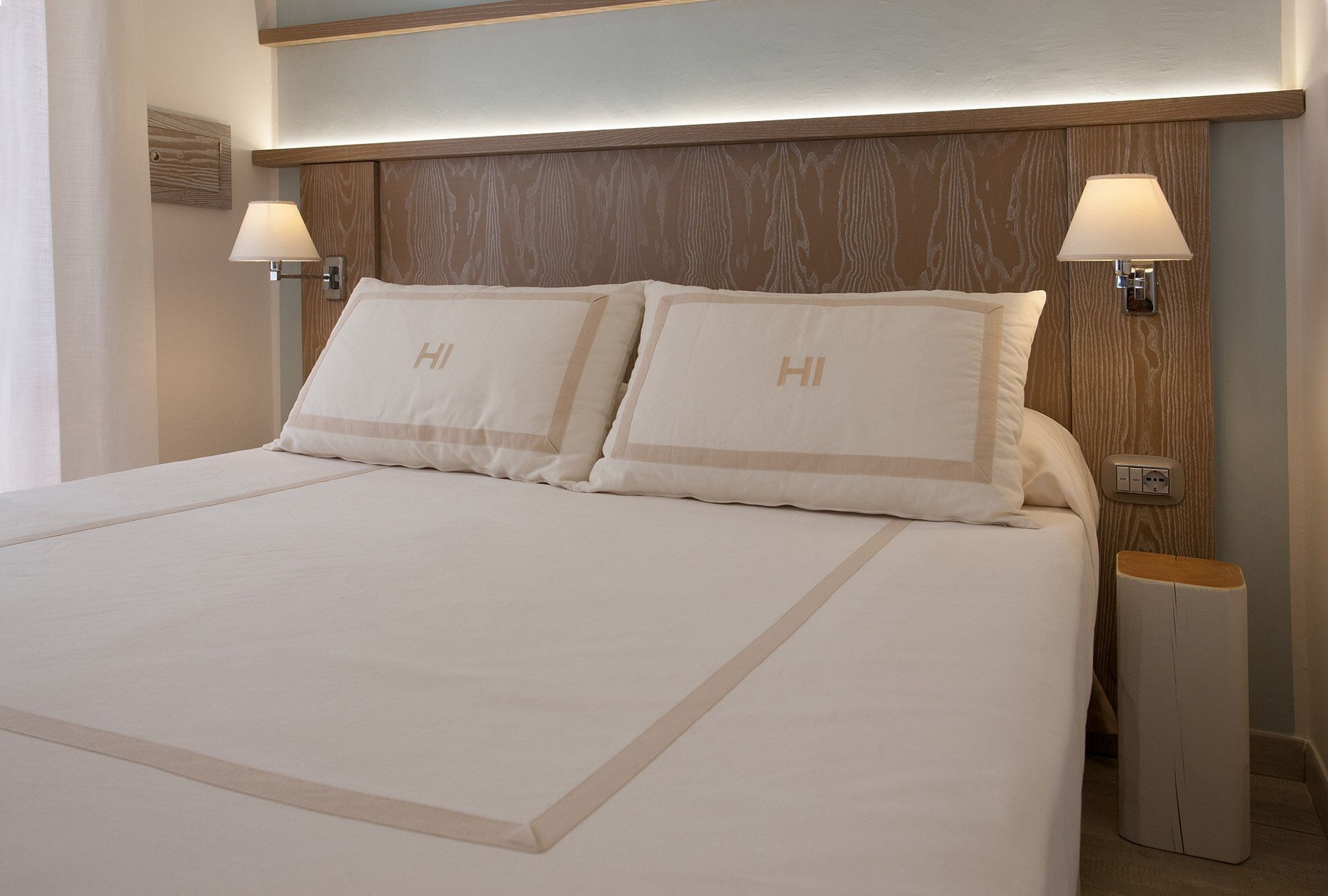 Quarto de hotel com uma cama grande, duas luminárias de cabeceira e uma cabeceira de madeira.