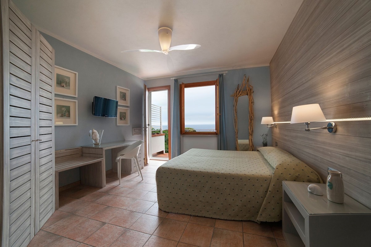 Ein Schlafzimmer mit Bett, Nachttisch, Schreibtisch und Stuhl. Ein Fenster bietet Blick aufs Meer.