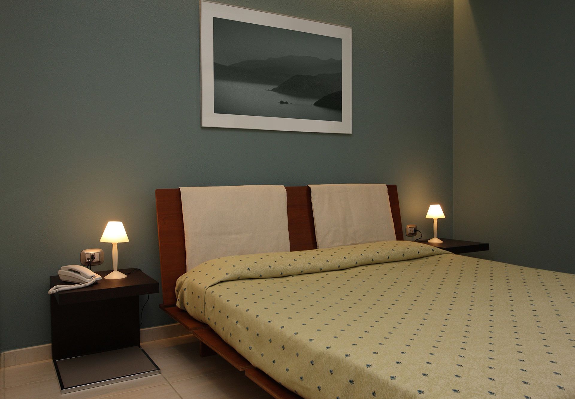 A bedroom with a bed, two bedside tables with lamps, a telephone, and a framed picture on the wall.