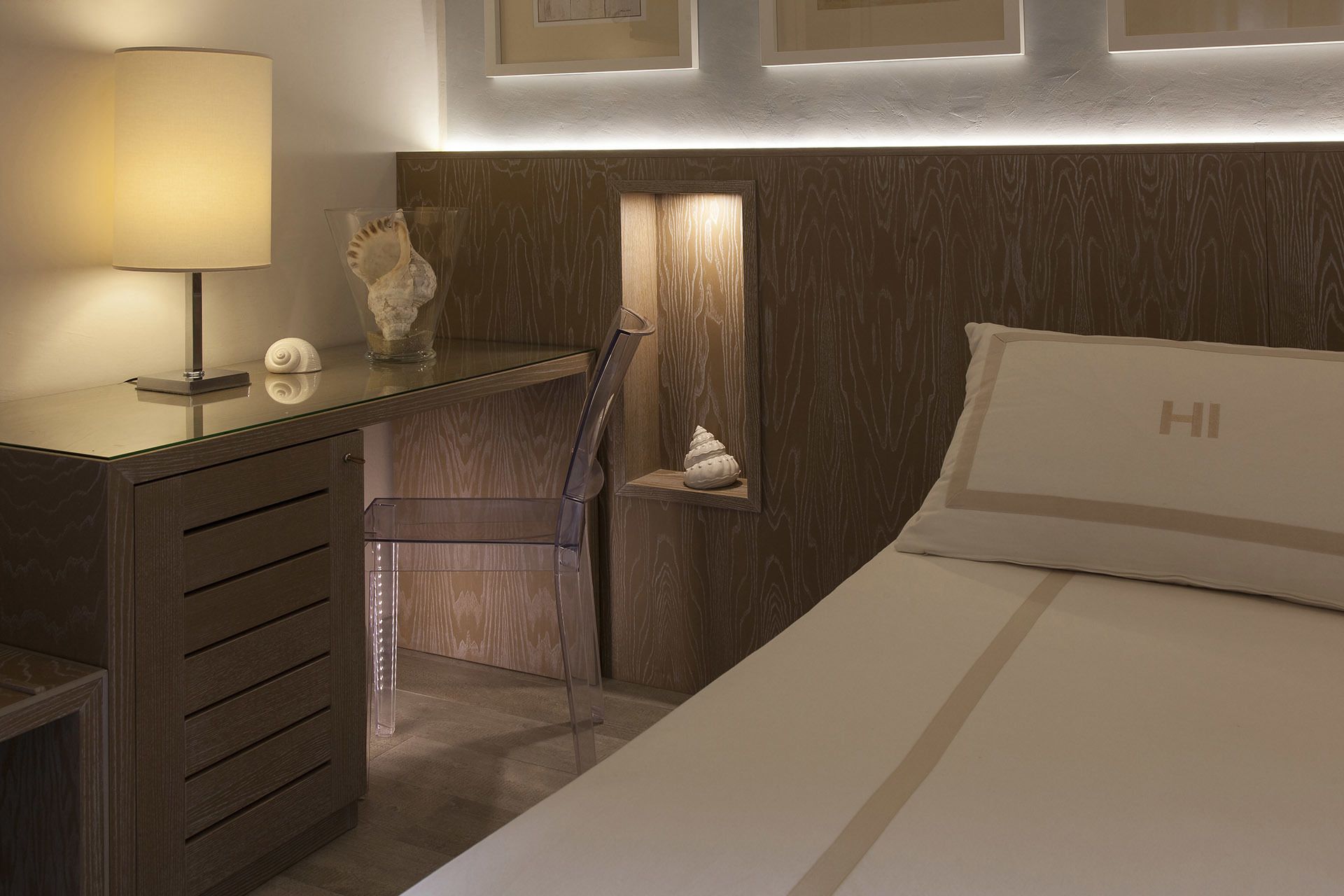 A hotel room with a wooden headboard, a desk with a lamp, a chair, and a bed with a pillow marked 'HI'.