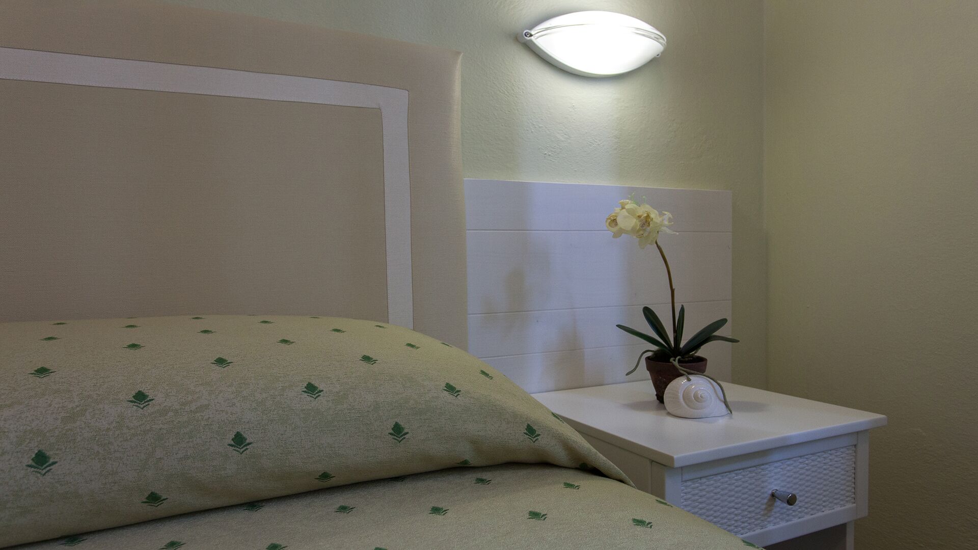 A hotel room with a bed, a nightstand with a potted plant, and a wall-mounted light fixture.