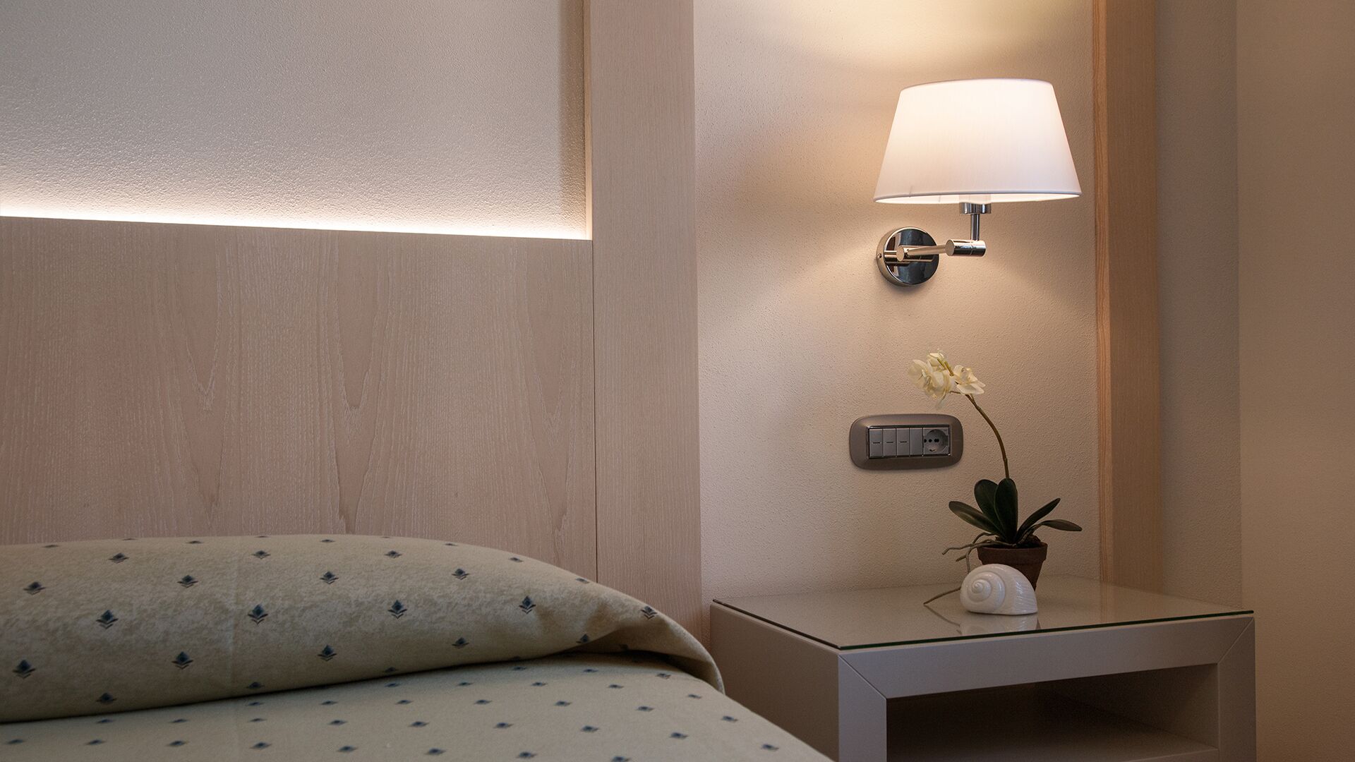 En pænt redt seng med pude med mønster, et sengebord med lampe, en lille urtepotte og en stikkontakt.