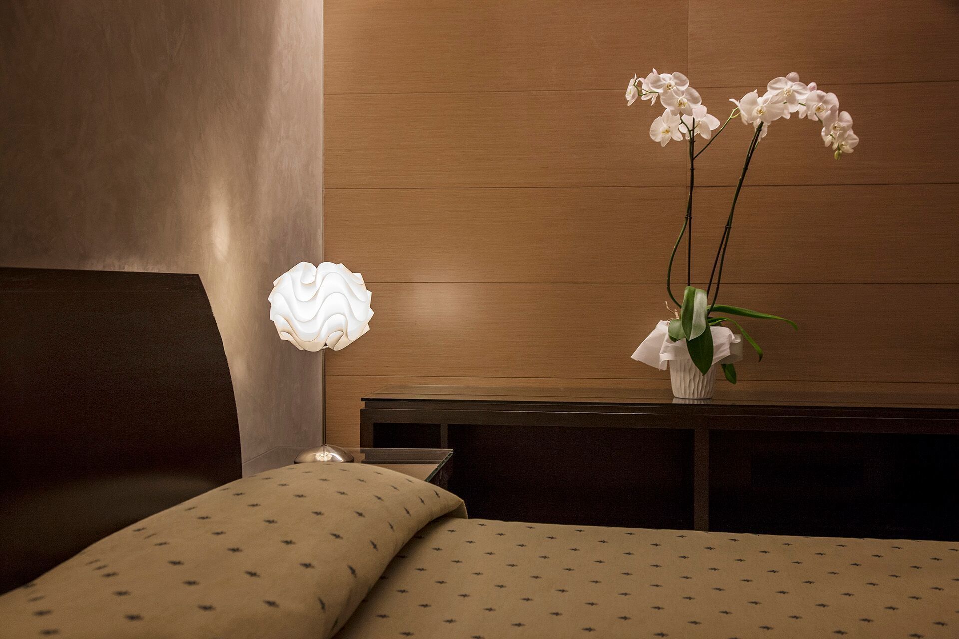 Une chambre à coucher avec une tête de lit en bois, une table de chevet surmontée d’une lampe blanche et un vase contenant des orchidées blanches.