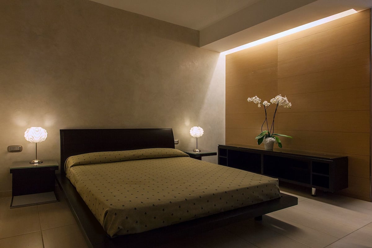Ein modernes Schlafzimmer mit einem Bett, zwei Nachttischlampen, einem Nachttisch, einem Holzkonsole und einer Topforchidee.